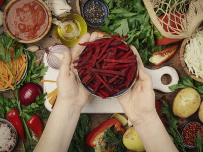 Ingredientes vegetarianos veganos frescos, verduras, especias, hierbas para una alimentación saludable en una mesa rústica de cocina de madera con tabla de cortar. Manos de mujer sosteniendo remolachas picadas. Concepto de cocina dietética, limpia y reconfortante
