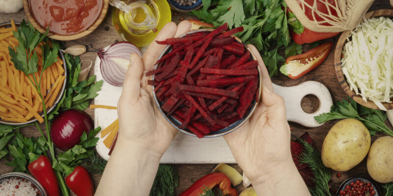 Ingredientes vegetarianos veganos frescos, verduras, especias, hierbas para una alimentación saludable en una mesa rústica de cocina de madera con tabla de cortar. Manos de mujer sosteniendo remolachas picadas. Concepto de cocina dietética, limpia y reconfortante