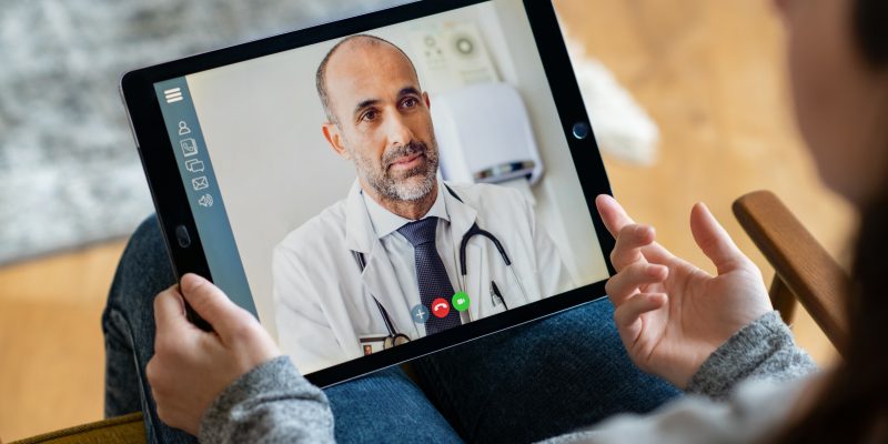 Vista posterior de una joven que hace una videollamada con su médico mientras se queda en casa. Primer plano de un paciente sentado en una videoconferencia de sillón con un médico general en una tableta digital. Niña enferma en consulta en línea con un médico maduro.