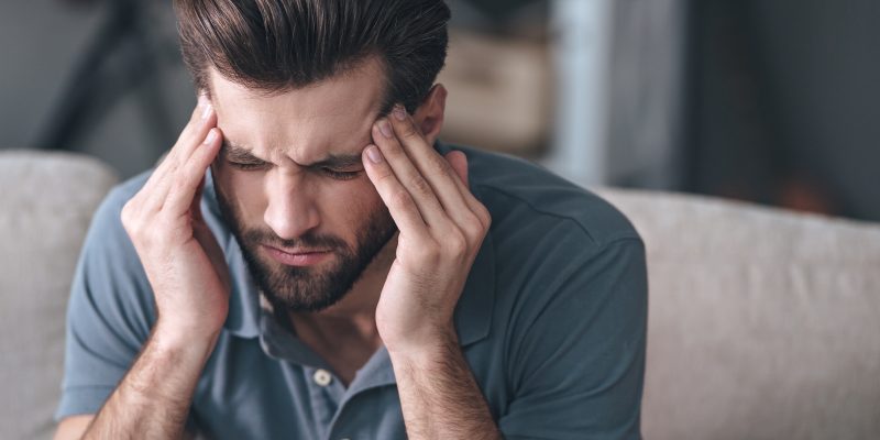 Sentirse estresado. Un joven apuesto y frustrado tocándose la cabeza y manteniendo los ojos cerrados mientras se sienta en el sofá de casa